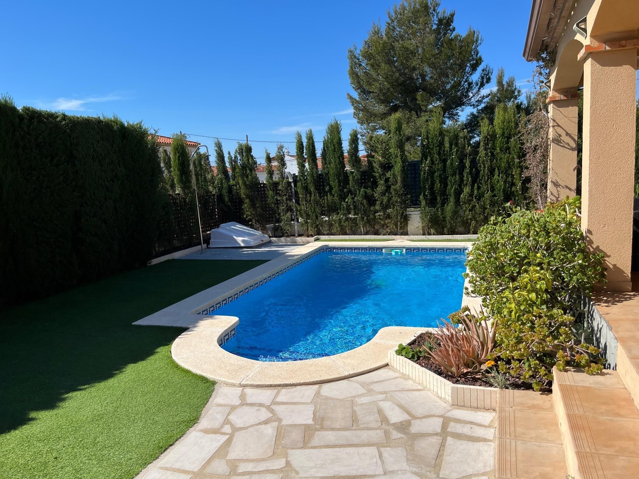 Une piscine dotée d’une plage en pierre, entourée d’une pelouse bien entretenue et de grands arbres.