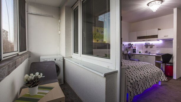 A compact room with a bed, a small table with a vase, a washing machine, and a kitchenette area.