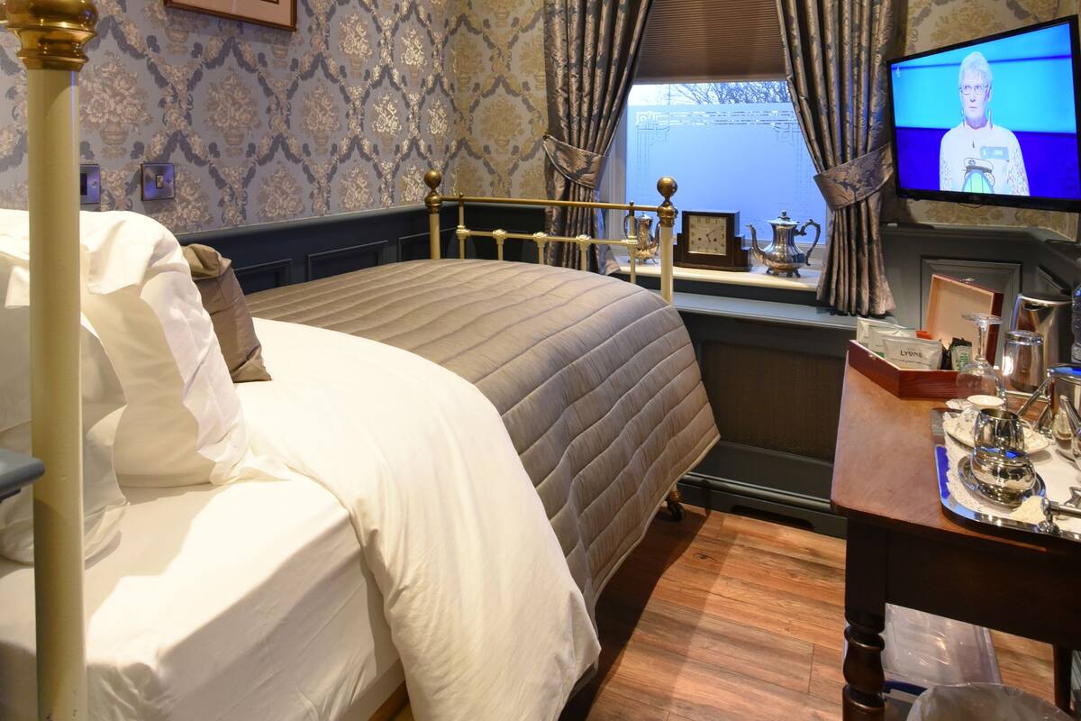 A hotel room with a bed, a television, a window with curtains, a small table with a lamp, and a picture on the wall.