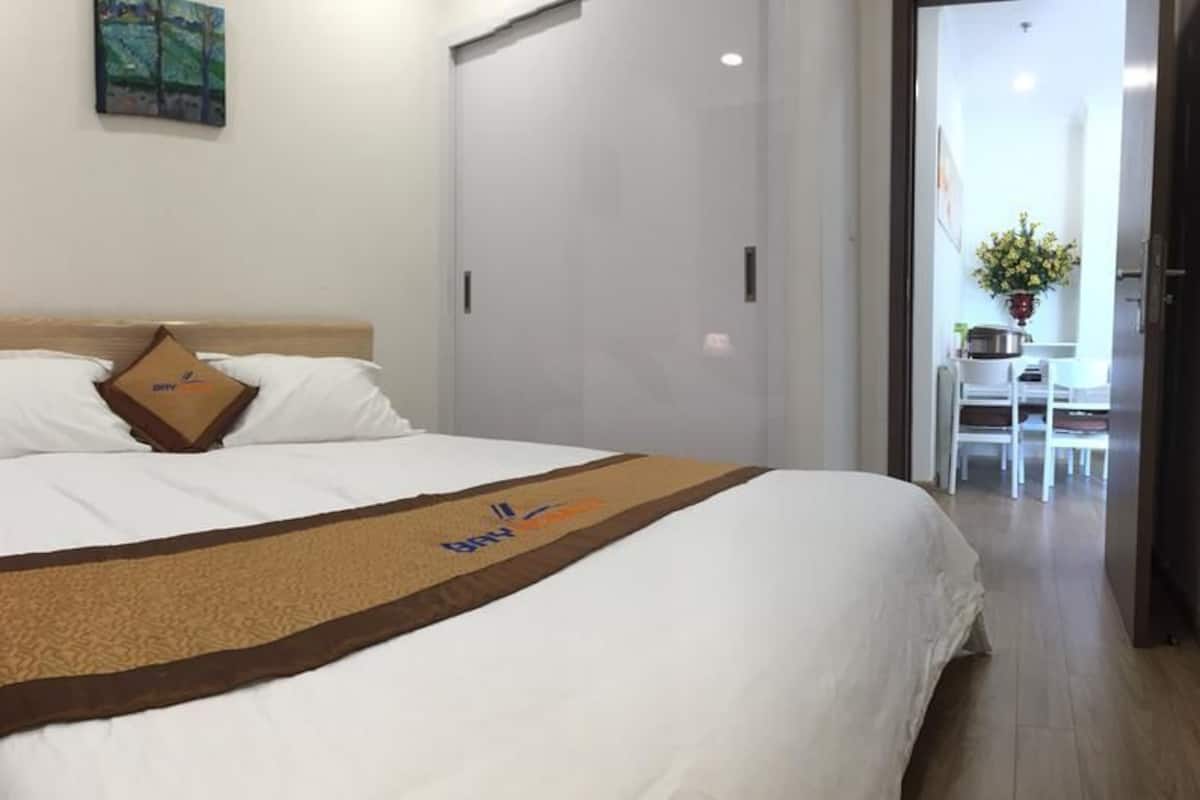 A hotel room with a bed, a nightstand with a lamp, a painting on the wall, and a view of a dining area with a table and chairs.