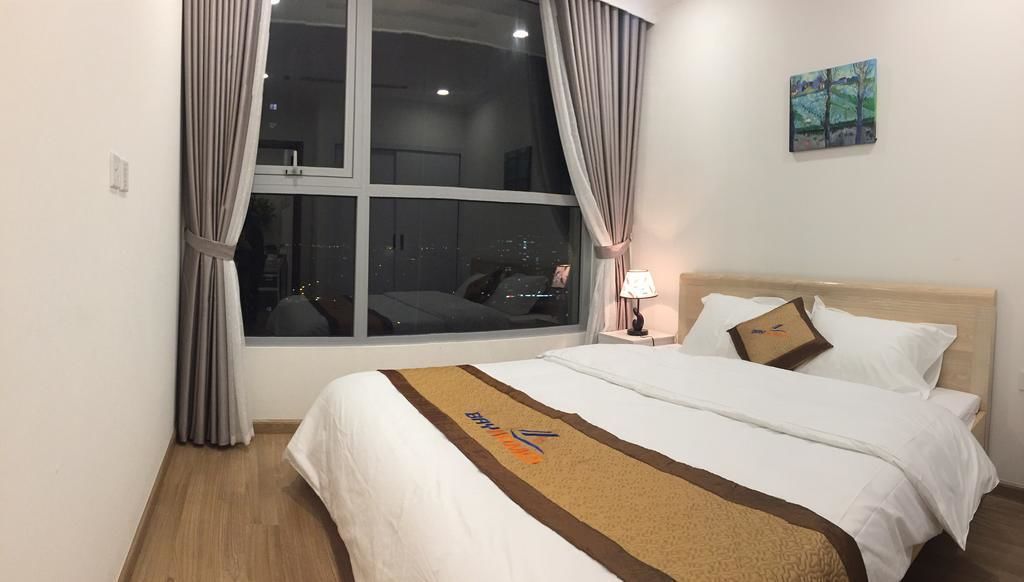 A hotel room with a large window, a bed with white bedding, a brown throw blanket, and a painting on the wall.