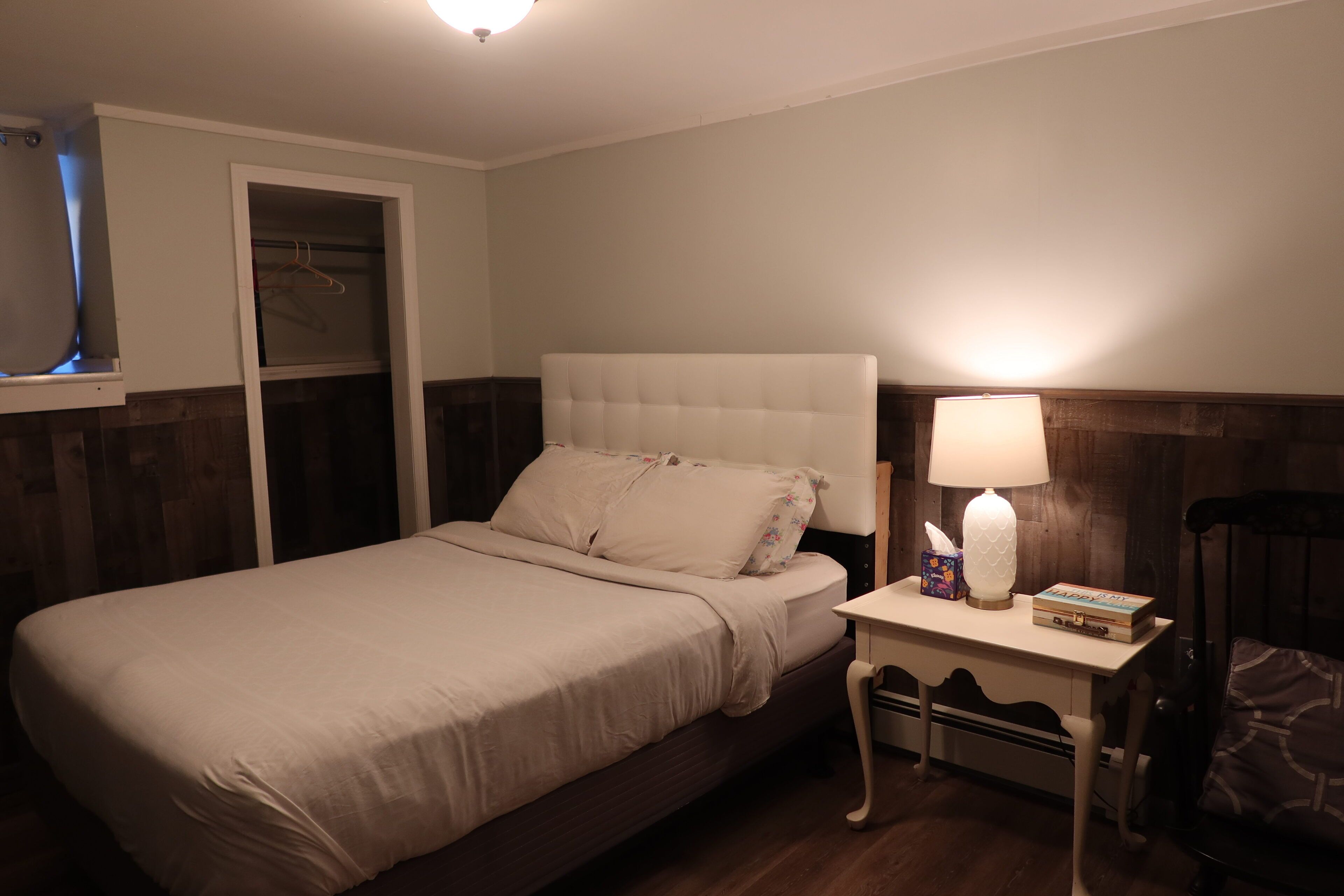 A neatly made bed with a white headboard, a bedside table with a lamp and a book, and a chair with a patterned cushion.