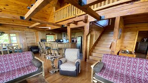 Interior d'una cabana de fusta amb una escala de fusta, una zona de menjador amb cadires i taula de fusta, i un saló amb sofàs i taula baixa.