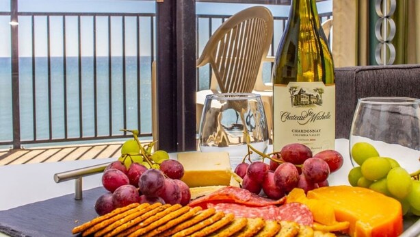 A table with a bottle of Chateau Michelle Chardonnay, grapes, cheese, and crackers on a balcony with a view of the sea. - Bring Your Family to Our Gorgeous Oceanfront Condo (Myrtle Beach)