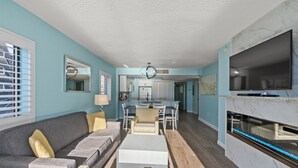 A modern living room with a sofa, coffee table, and TV. There is a kitchen in the background with a dining area. - Bring Your Family to Our Gorgeous Oceanfront Condo (Myrtle Beach)