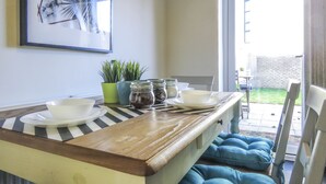 Ein Essbereich mit einem Holztisch, gestreiften Platzsets und blauen, gepolsterten Stühlen. Üchtöpfe, Blumentöpfe und ein gerahmtes Bild sind an der Wand zu sehen.