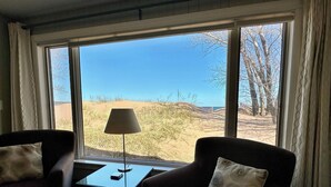 A room with two armchairs, a small table, and a lamp. A large window offers a view of sand dunes and trees. - Beachfront Getaway on Park Point - Easy Walking Distance to Canal Park (Duluth)