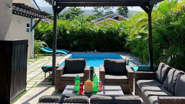 Un patio exterior con mobiliario de mimbre, una piscina y una vista de una casa al fondo.