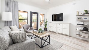 A modern living room with a sofa, coffee table, and TV. There is a balcony with a view of greenery. - Ultra-Clean, Designer Condo. Walk to grocery, restaurants, coffee, and shopping! (Scottsdale)