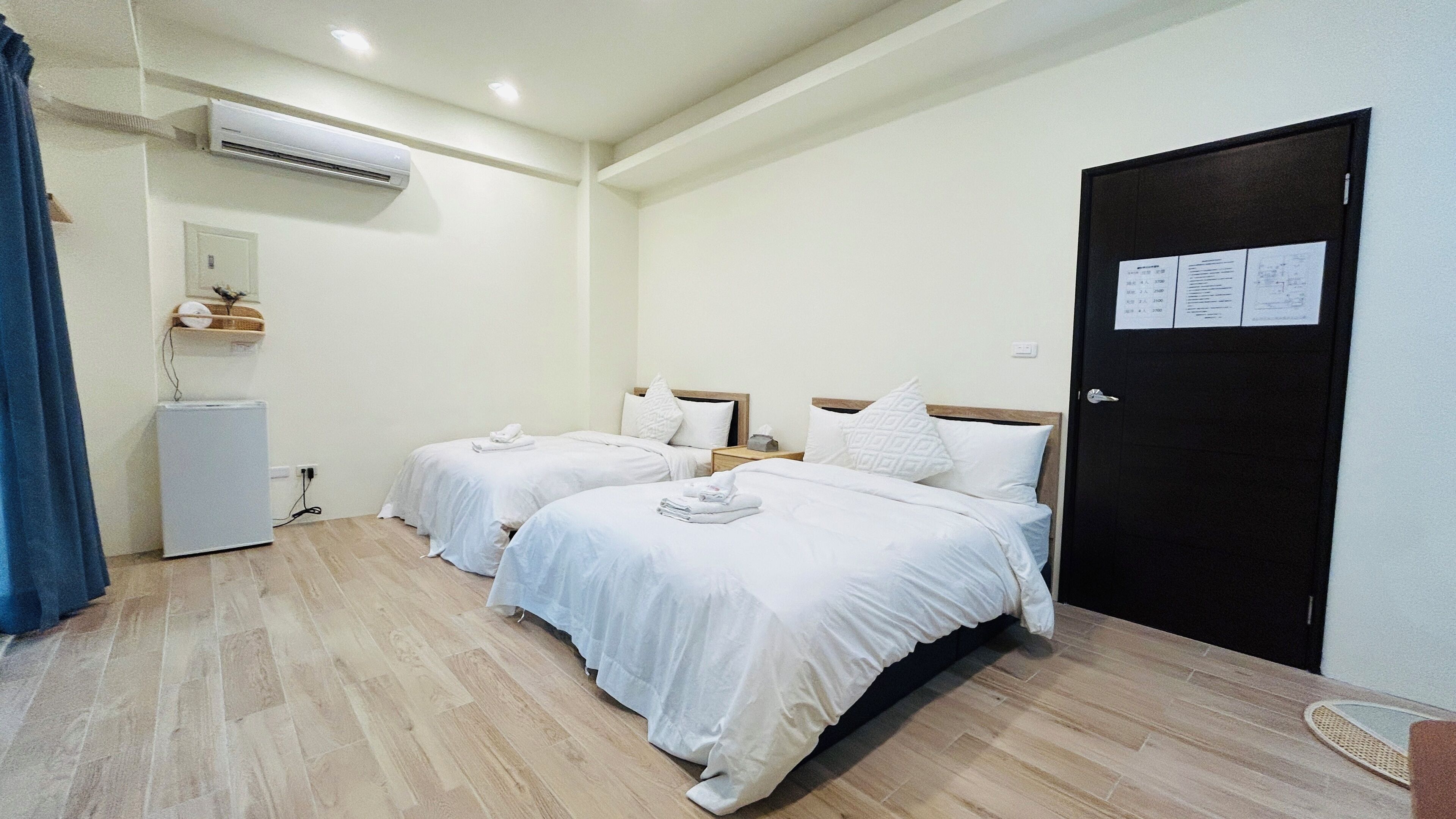 A hotel room with two beds, a small refrigerator, an air conditioning unit, and a door with a notice.