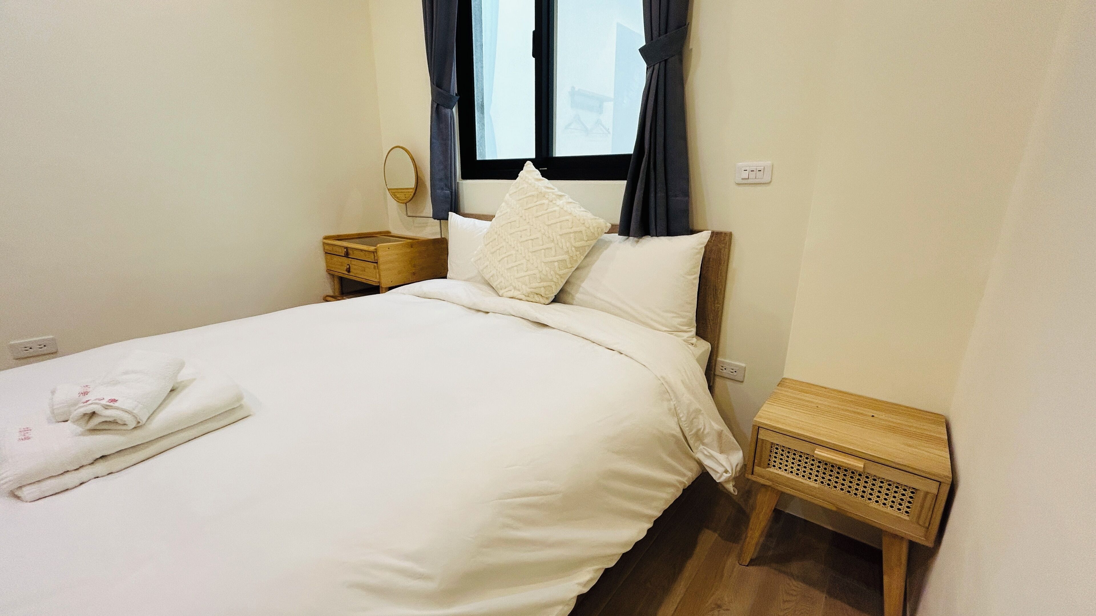 A neatly made bed with white linens and a folded towel, a bedside table with a mirror, a wooden nightstand, and a window with curtains.