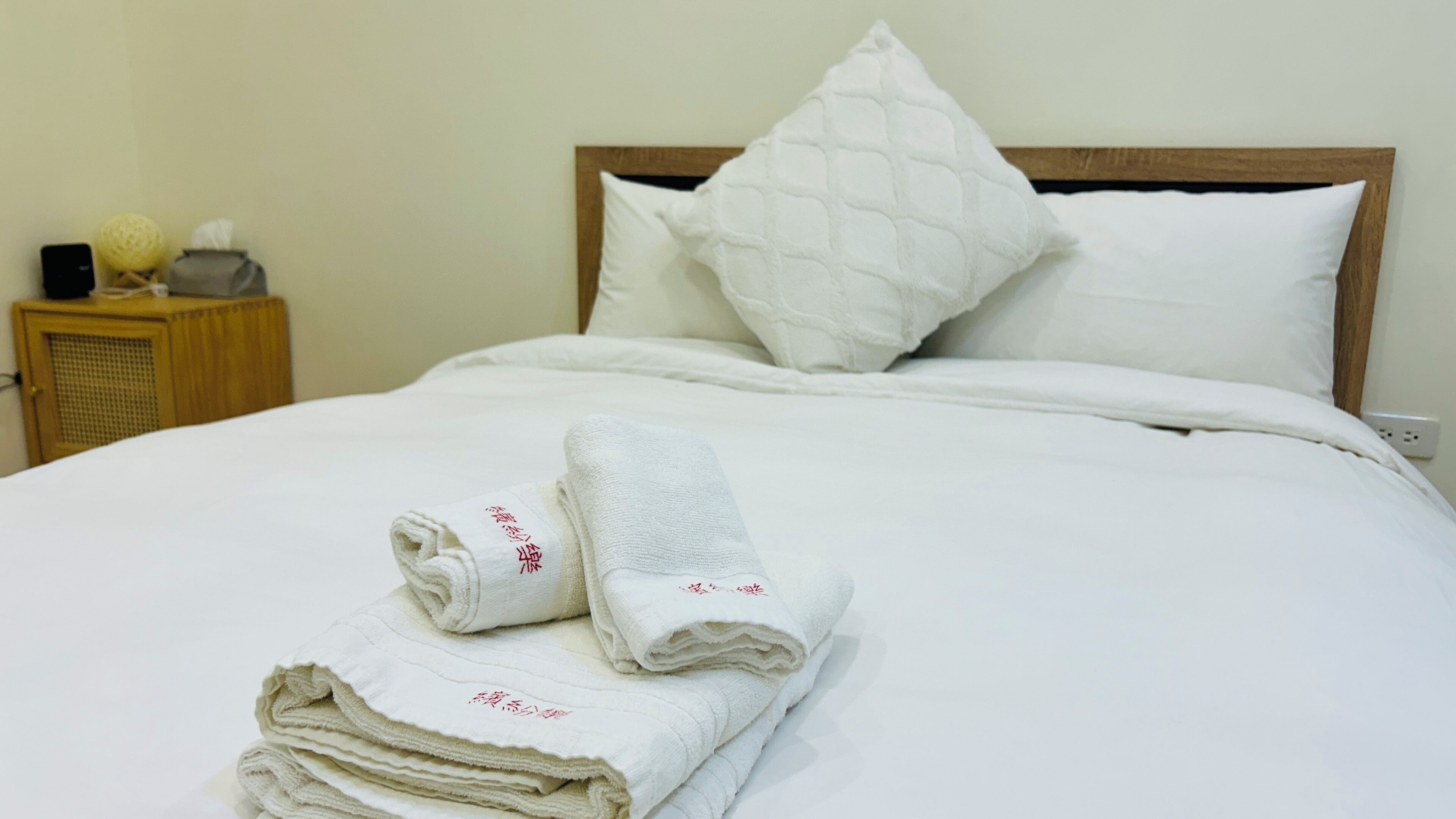 A neatly made bed with white linens and pillows, a wooden nightstand with a lamp and a small decorative item, and a folded towel with embroidered text.