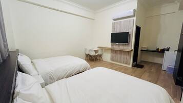A hotel room with two single beds, a small desk with a chair, a television mounted on the wall, and a refrigerator.