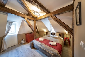 A bedroom with a sloped ceiling, wooden beams, a bed with a red blanket, two bedside tables with lamps, and a window with curtains. - OLD TOWN -2 BEDROOM 2 BATHROOM max 8 pers (Prague 1)