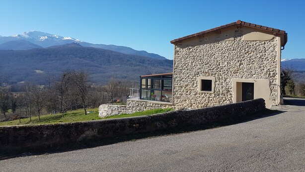 A stone building with a small window and a glass door, set against a backdrop of mountains and clear sky. - The barn corner of paradise (Leychert)