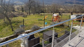 A view from a balcony overlooking a grassy area with playground equipment, a fenced play area, and a mountain range in the background. - The barn corner of paradise (Leychert)