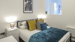 A modern bedroom with a bed, bedside lamps, a framed picture, and a window with a view. - Tudors eSuites Birmingham Apartments Jewellery Quarter (Birmingham)