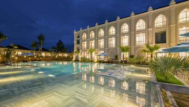 Una amplia piscina bien iluminada con sillas de descanso y sombrillas, rodeada por un edificio de estilo clásico con ventanales arqueados.