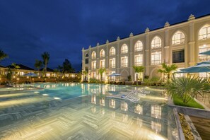A large, well-lit swimming pool with lounge chairs and umbrellas, surrounded by a building with arched windows and a classic architectural style. - My Son Heritage Resort And Spa (Da Nang)
