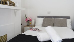 Un lit bien fait, avec du linge de lit blanc et une serviette roulée, une table de chevet ornée d’un vase de fleurs et d’un magazine, et une étagère avec des objets décoratifs, notamment une plante et des figurines.