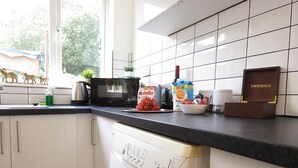 Une cuisine avec des meubles blancs, un plan de travail noir, un four à micro-ondes, un grille-pain, un pot de Nutella, un bol de raisins, un carton de jus d’orange et une boîte en bois étiquetée « TWININGS ».