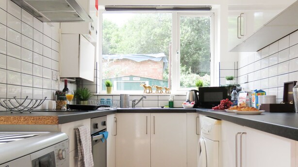 Une cuisine compacte avec des meubles blancs, un four encastré, un lave-linge et une fenêtre donnant sur un espace verdoyant et un bâtiment en briques.