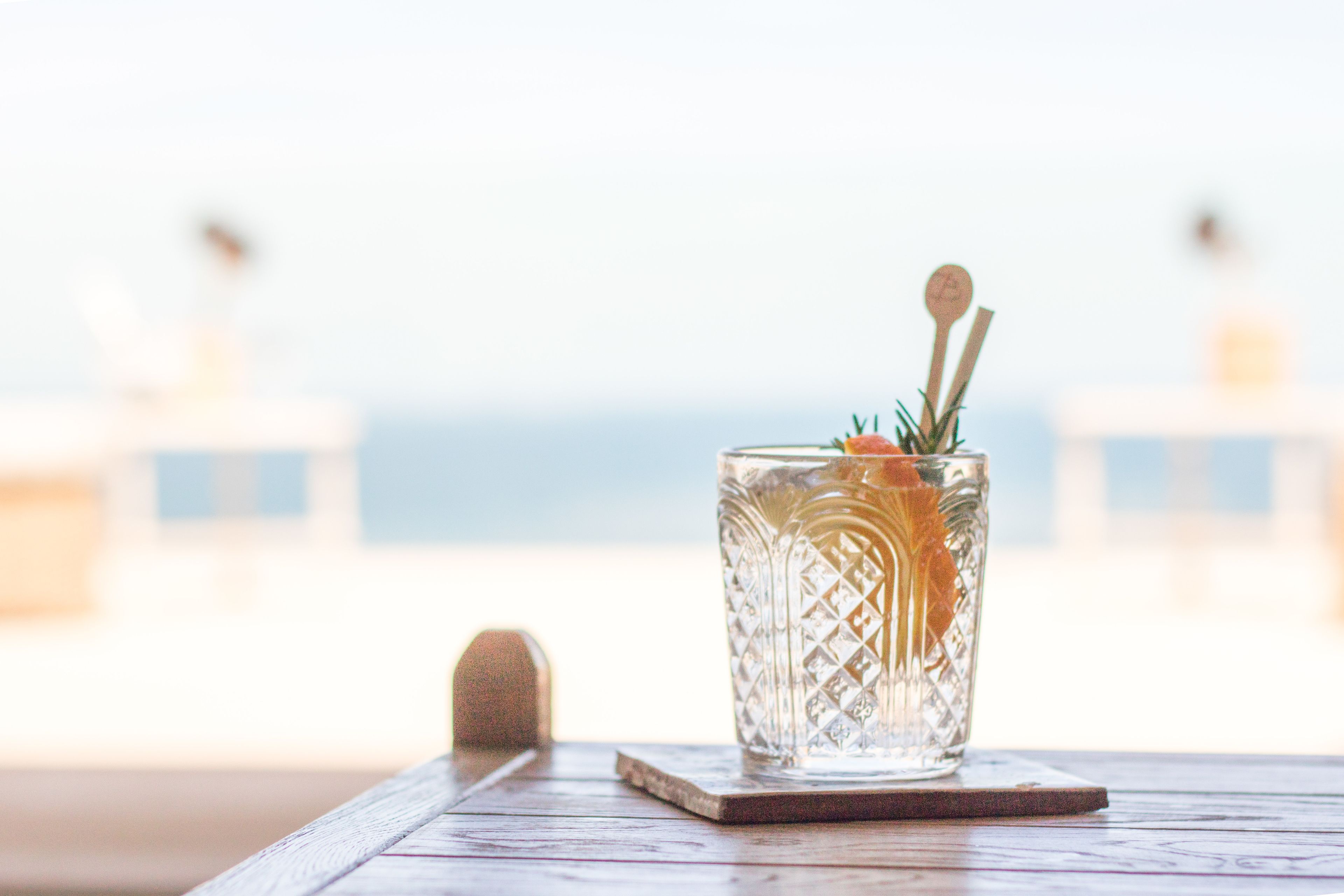 A clear glass with a straw and a wooden spoon on a coaster, placed on a wooden table with a blurred background.