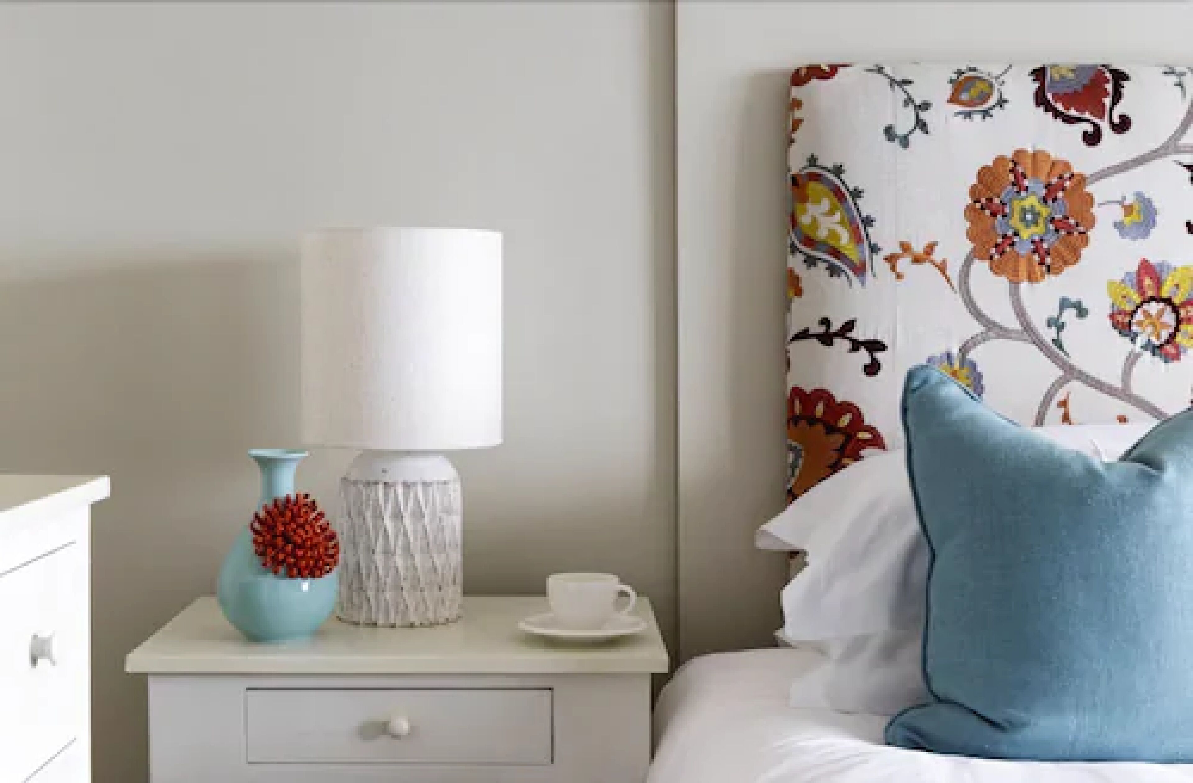 En pænt redt seng med en blå pude, et hvidt natbord med en lampe, en vase med røde blomster og en kop på bordet.