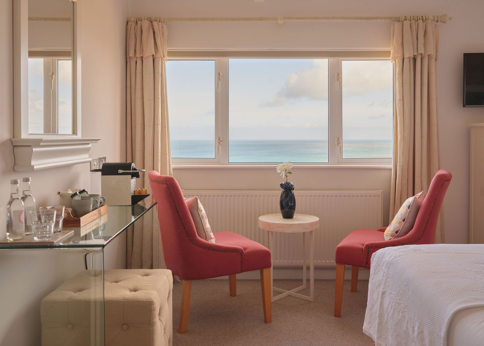 A hotel room with a view of the sea, two red chairs, a small round table with a vase, and a bed with a white cover.