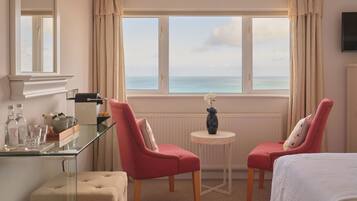 A hotel room with a view of the sea, two red chairs, a small round table with a vase, and a bed with a white cover.