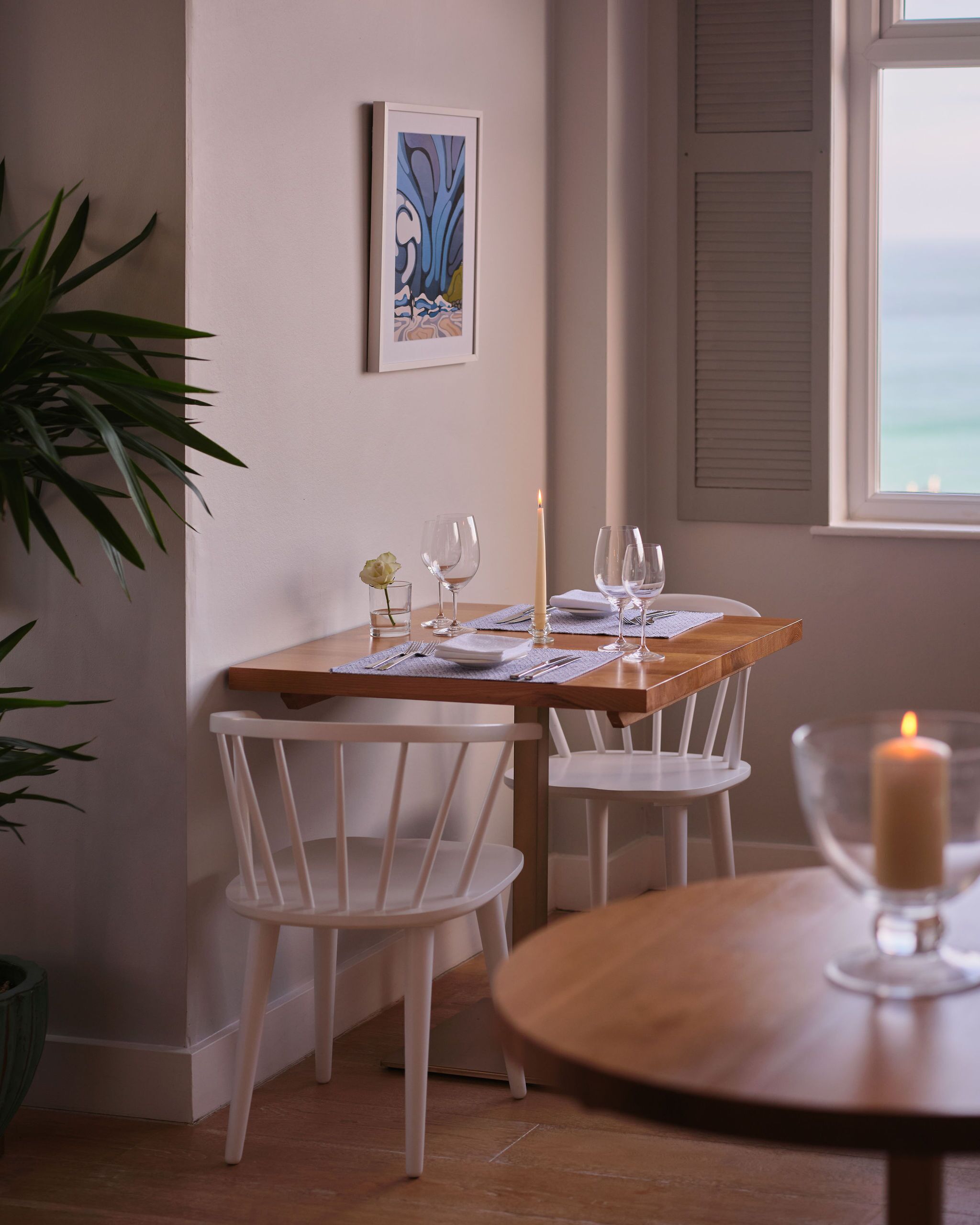 A dining area with a wooden table set for two, white chairs, a candle, and a framed artwork on the wall.