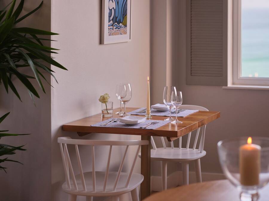 A dining area with a wooden table set for two, white chairs, a candle, and a framed artwork on the wall.