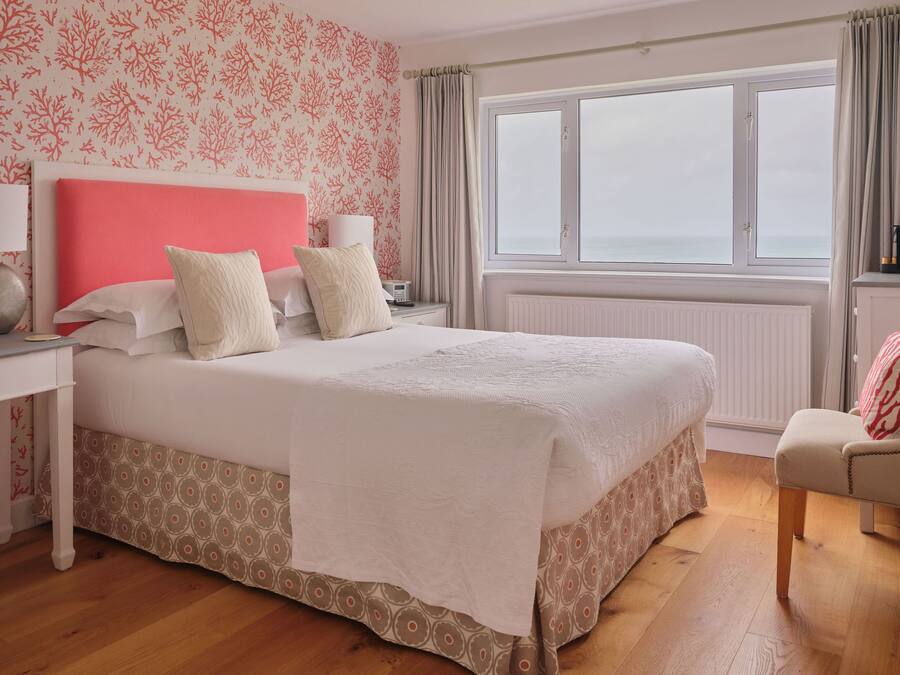 Habitación de hotel con una cama grande, una mesita de noche, una silla, un ventanal con vistas y un papel tapiz estampado.
