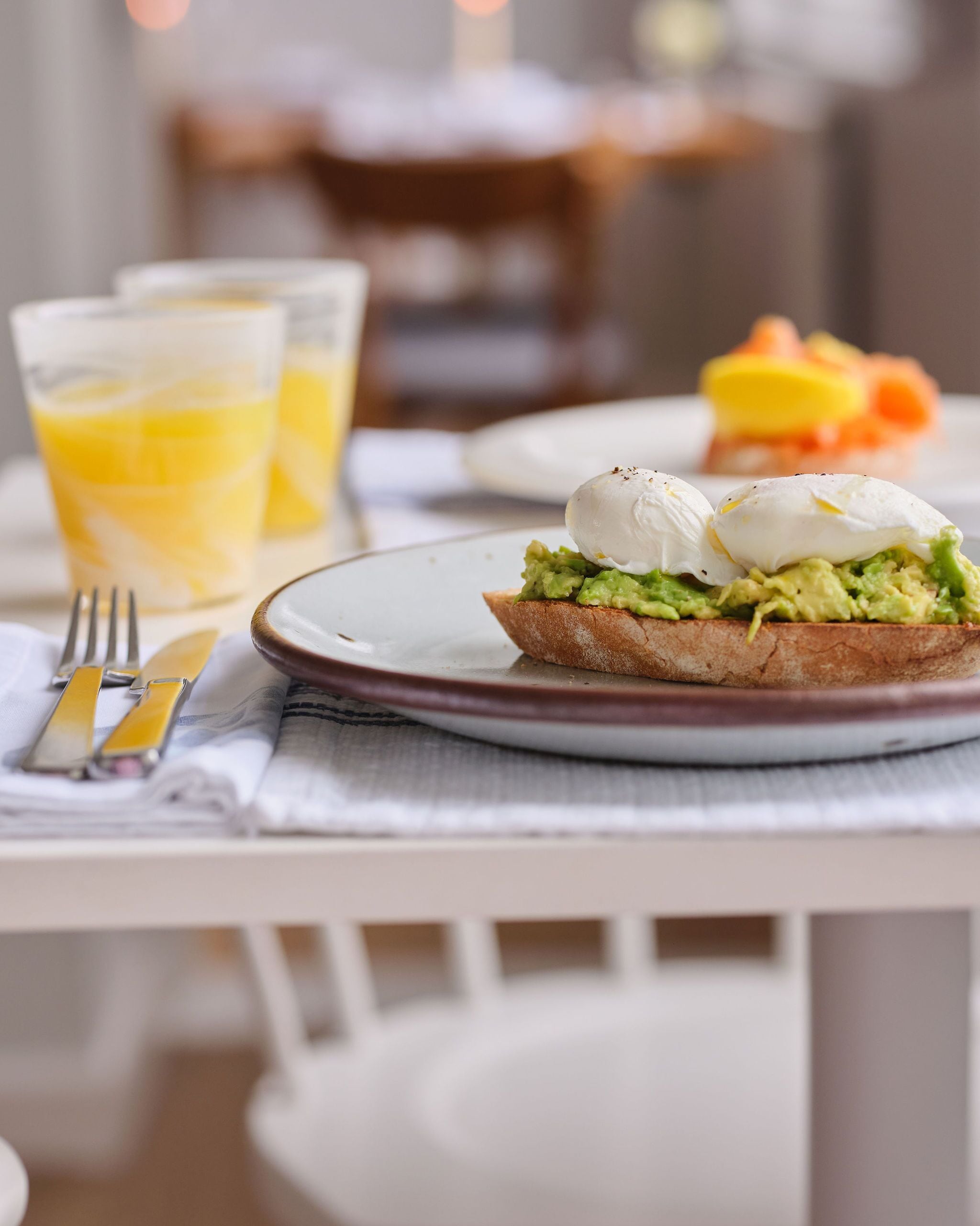 A breakfast setting with a plate of avocado toast topped with poached eggs, a glass of orange juice, and a bowl of fruit.