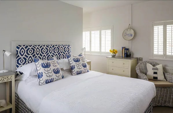 A bedroom with a large bed, a bedside table with a lamp, a dresser, a chair, and a wall clock. - Boskerris Hotel (St Ives)