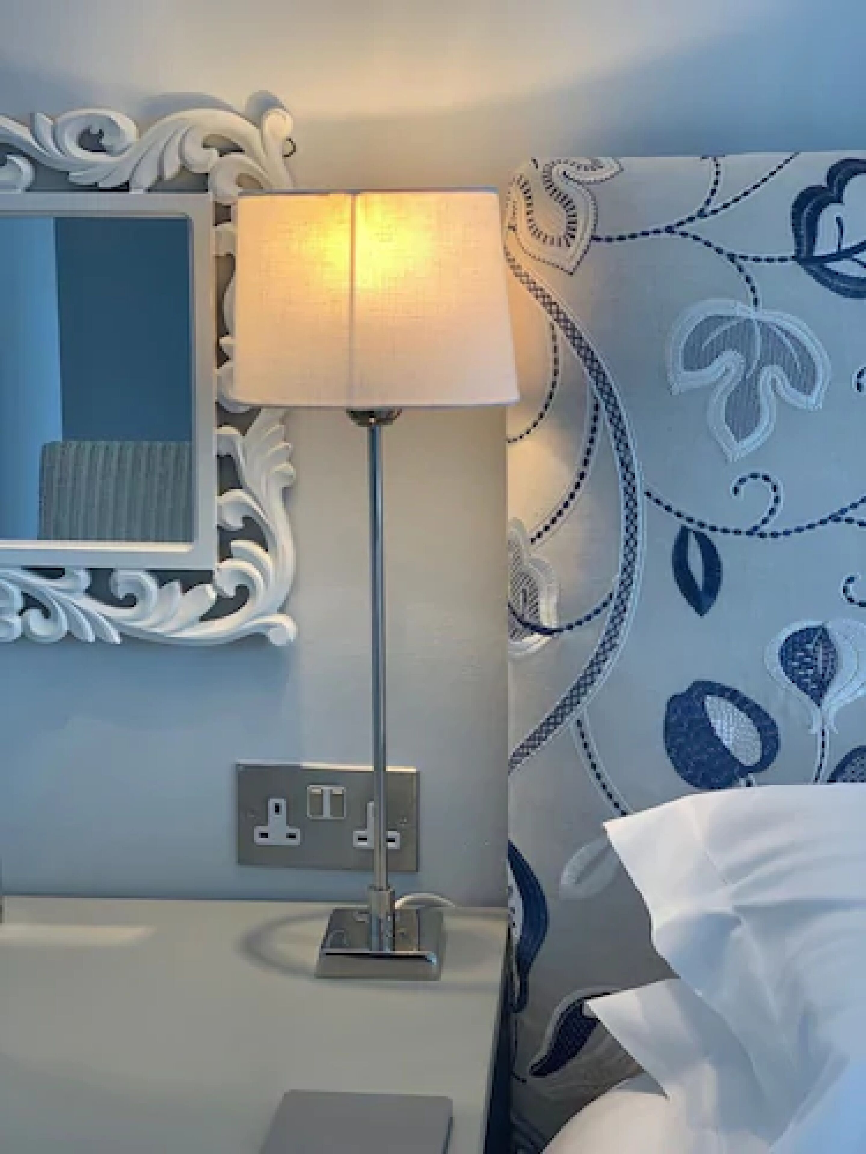 Habitación de hotel ordenada con una mesita de noche, una lámpara, un enchufe y un papel tapiz estampado.