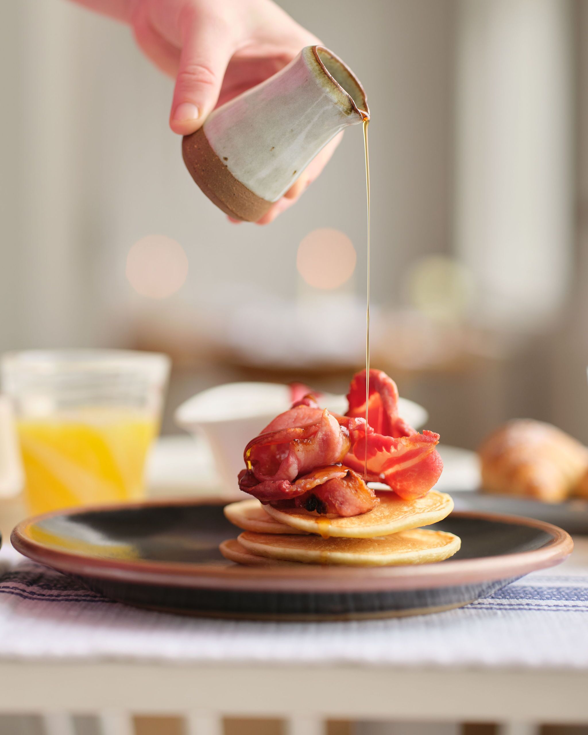 A breakfast plate with pancakes topped with bacon and syrup being poured from a pitcher.