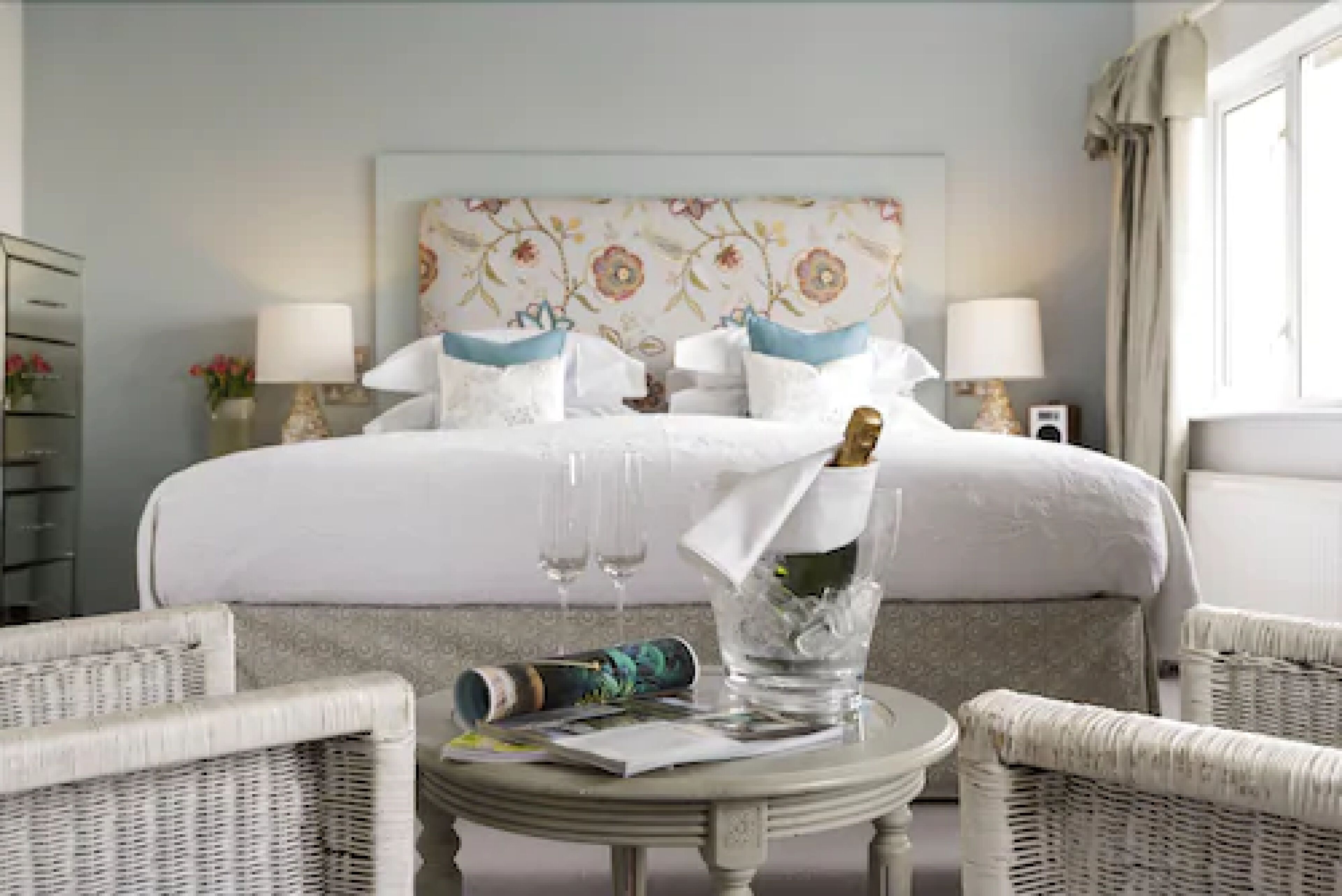 A hotel room with a bed, two bedside lamps, a floral headboard, a wicker chair, a small round table with a bottle and glasses, and a window with curtains.