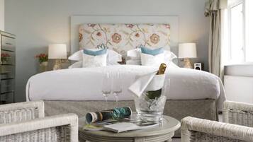 A hotel room with a bed, two bedside lamps, a floral headboard, a wicker chair, a small round table with a bottle and glasses, and a window with curtains.