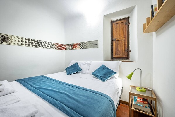 Une chambre à coucher avec un lit, des oreillers, une couverture, une étagère en bois avec des livres, une lampe et une œuvre d’art accrochée au mur.