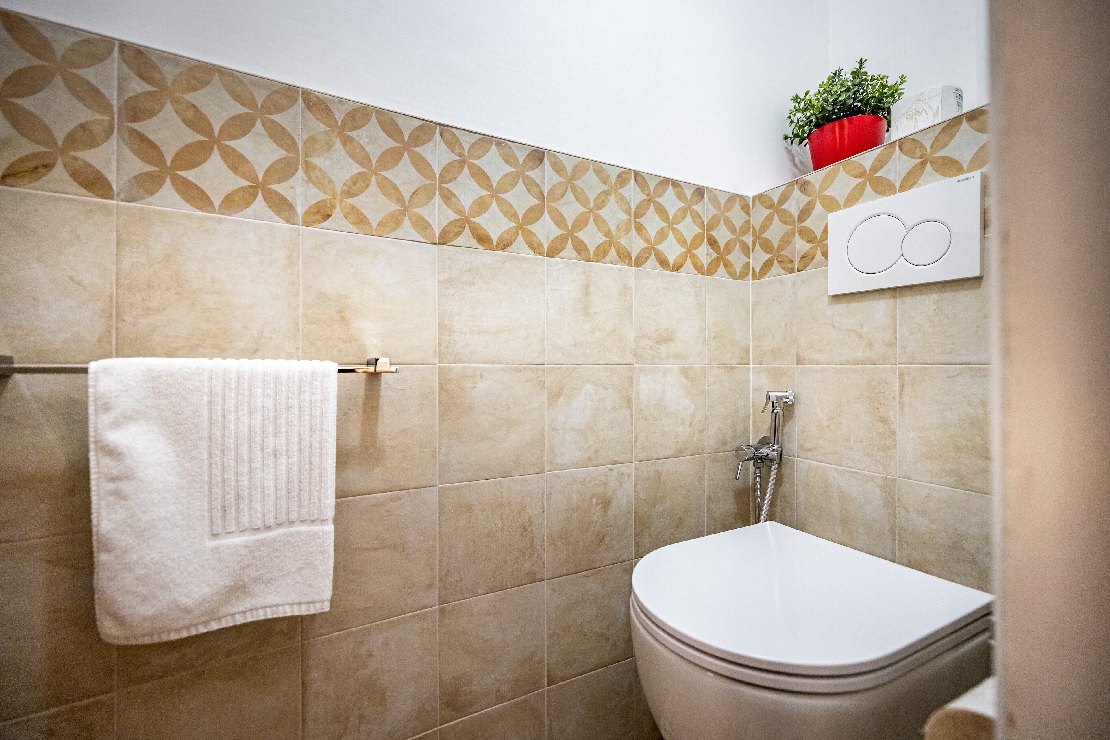 Un baño con baldosas color beige, una toalla blanca en un perchero, un inodoro y una planta en maceta en la pared.