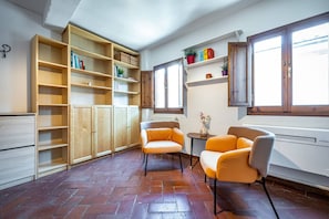 Una habitación con suelo de baldosas, estanterías de madera, dos sillones, un radiador y una ventana.