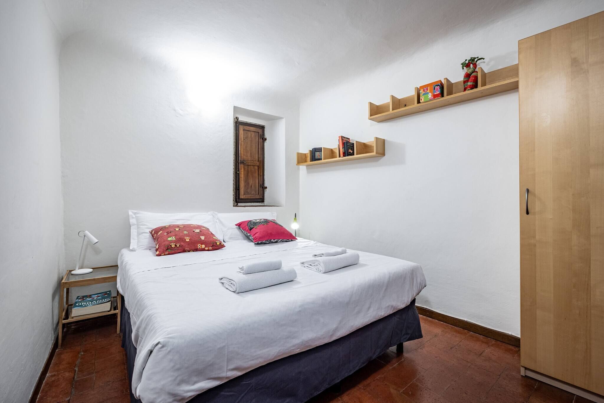 A bedroom with a bed, pillows, a nightstand, a shelf with books, a wardrobe, and a door.