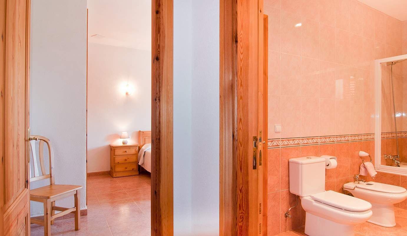 A bathroom with a toilet, sink, and shower. There is a wooden chair and a bedside table with a lamp in the adjacent room.