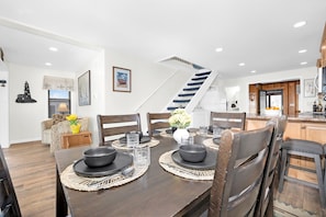 Comedor con mesa de madera dispuesta para seis personas, una escalera y una zona de cocina con armarios de madera.