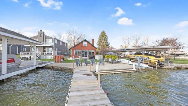 A wooden dock leading to a marina with boats, a red house, and a covered seating area.