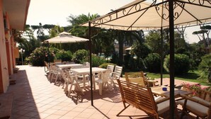 An outdoor patio with tiled flooring, white plastic chairs, and tables, surrounded by lush greenery and palm trees. - Hotel Parco dei Principi (Anzio)