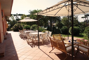 Eine Terrasse im Freien mit gefliestem Boden, weißen Plastikstühlen und Tischen, umgeben von üppiger Vegetation und Palmen.