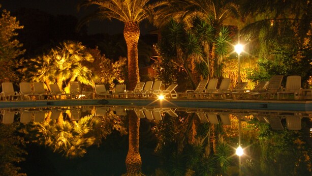 A nighttime pool area with lounge chairs and palm trees, reflecting in the water. - Hotel Parco dei Principi (Anzio)