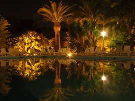 Ein Poolbereich bei Nacht mit Liegestühlen und Palmen, deren Spiegelung sich im Wasser zeigt.